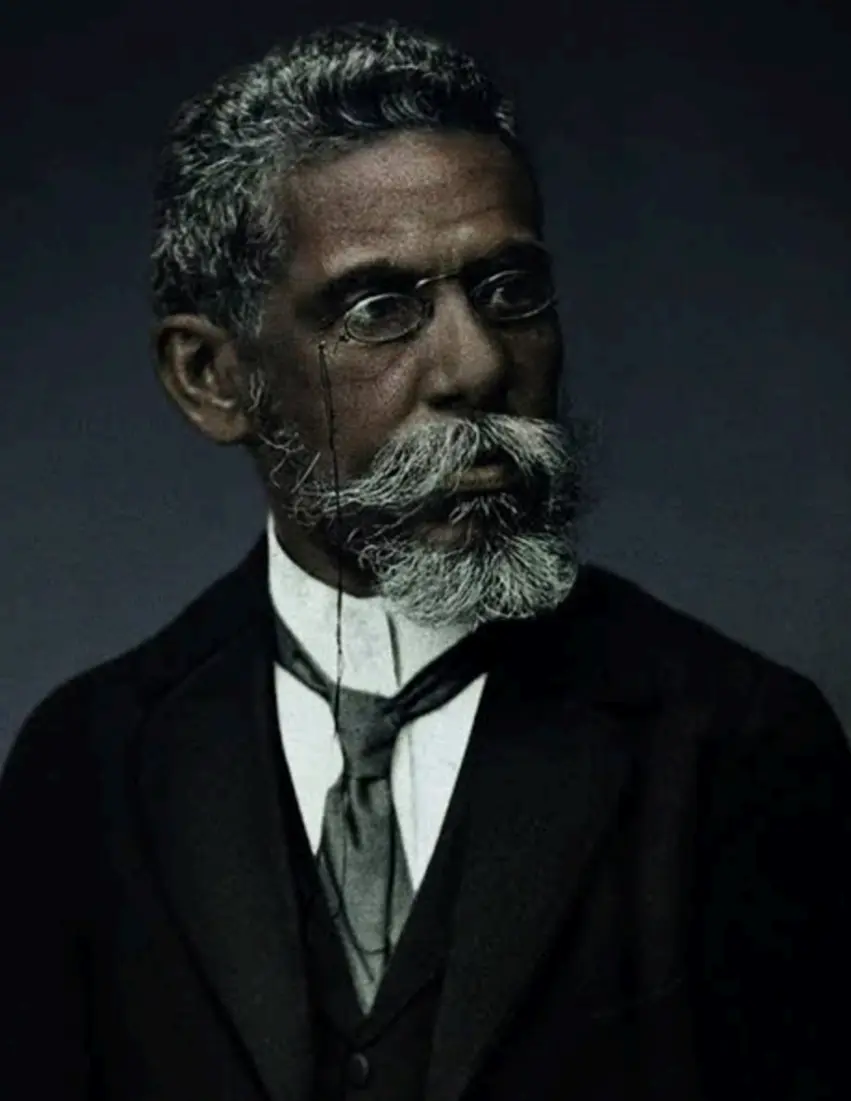 Homem adulto em terno preto, camisa branca e gravata preta, cabelos grisalhos e barba densa, óculos finos, fundo escuro.