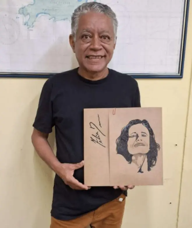Homem sorridente com livro artesanal, parede bege e mapa mundial ao fundo.