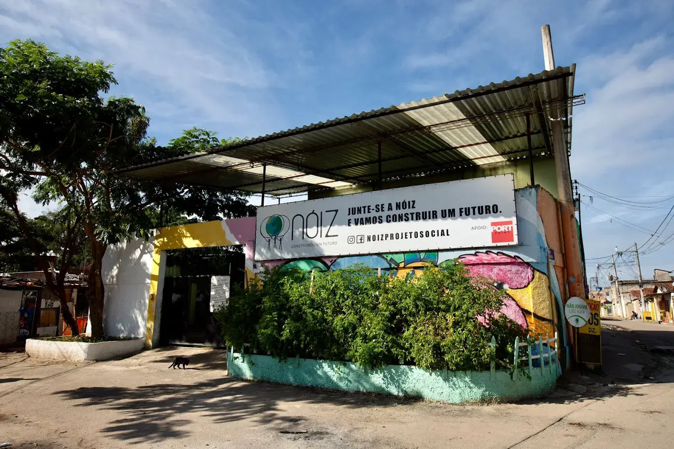 Um local com estrutura metálica e telhado verde, árvore grande à esquerda, construção colorida no centro, placa "Nóiz" e mensagem social à direita, céu azul com nuvens.
