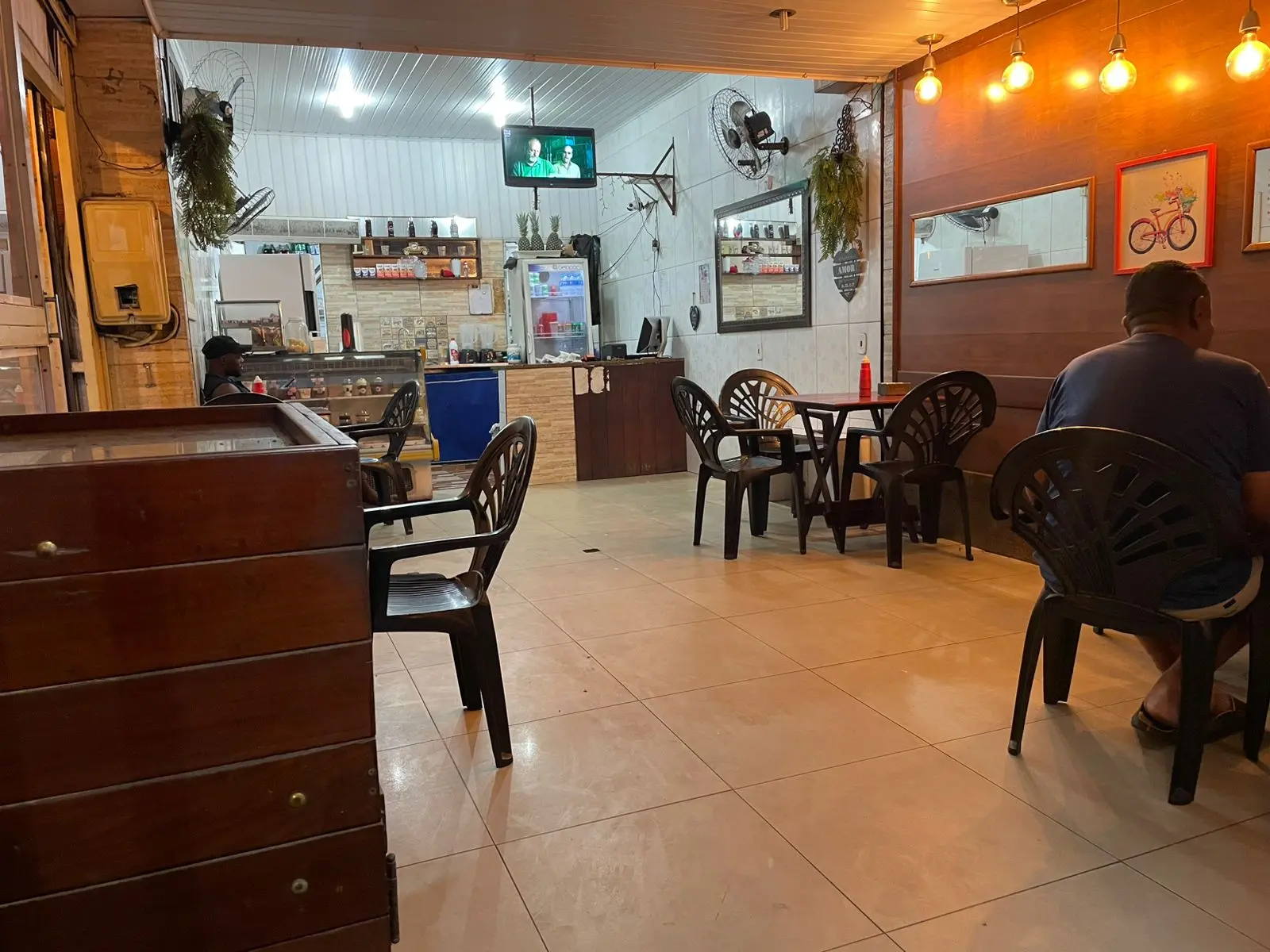 Sala de lanchonete com mesas, cadeiras, caixa de madeira, barra com geladeira e prateleiras, parede de madeira com quadros e luminárias, e televisão exibindo cena.