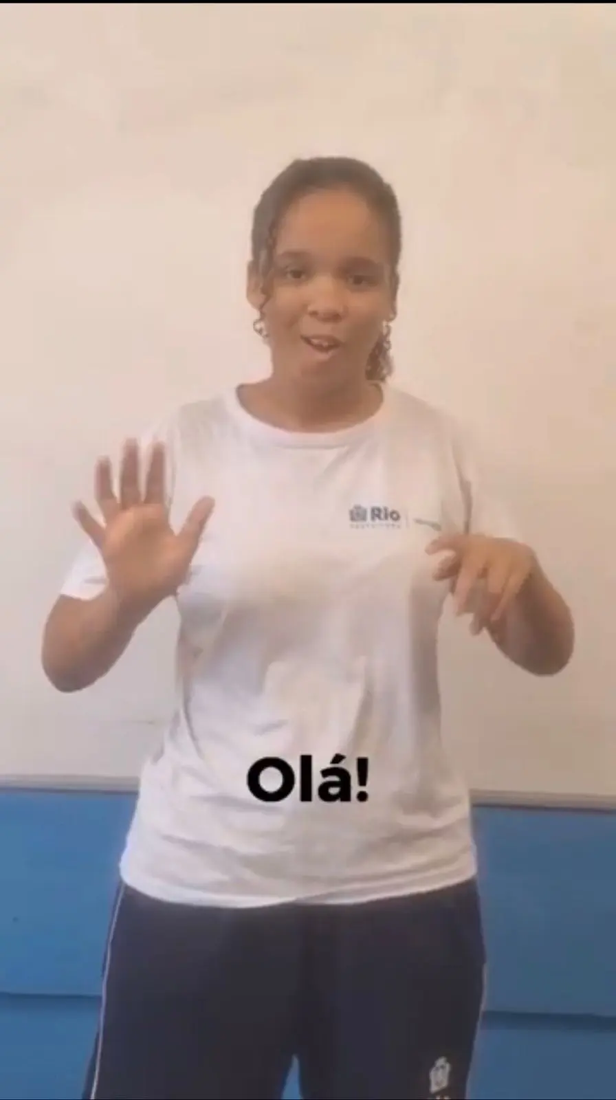 Pessoa sorrindo em ambiente interno, vestindo camiseta branca com logotipo "Rio" e texto "Olá!".