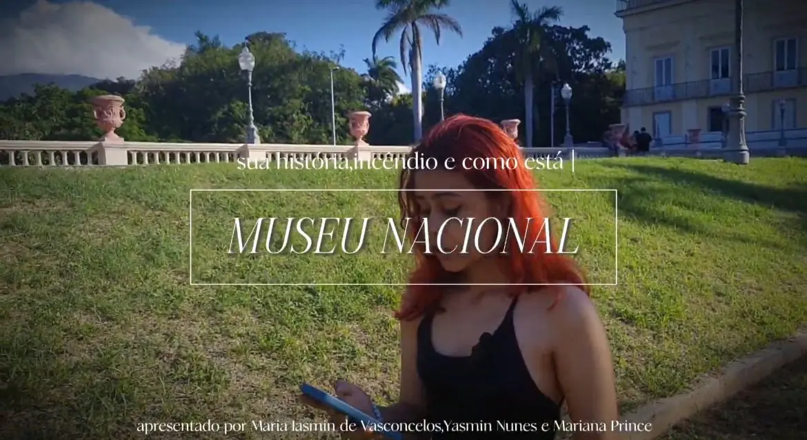 Parque com edifício histórico, pessoa com cabelos vermelhos sentada no gramado usando celular.