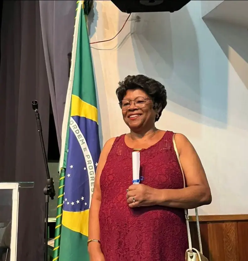 Mulher sorridente com vestido marrom escuro, segurando diploma e bandeira brasileira.