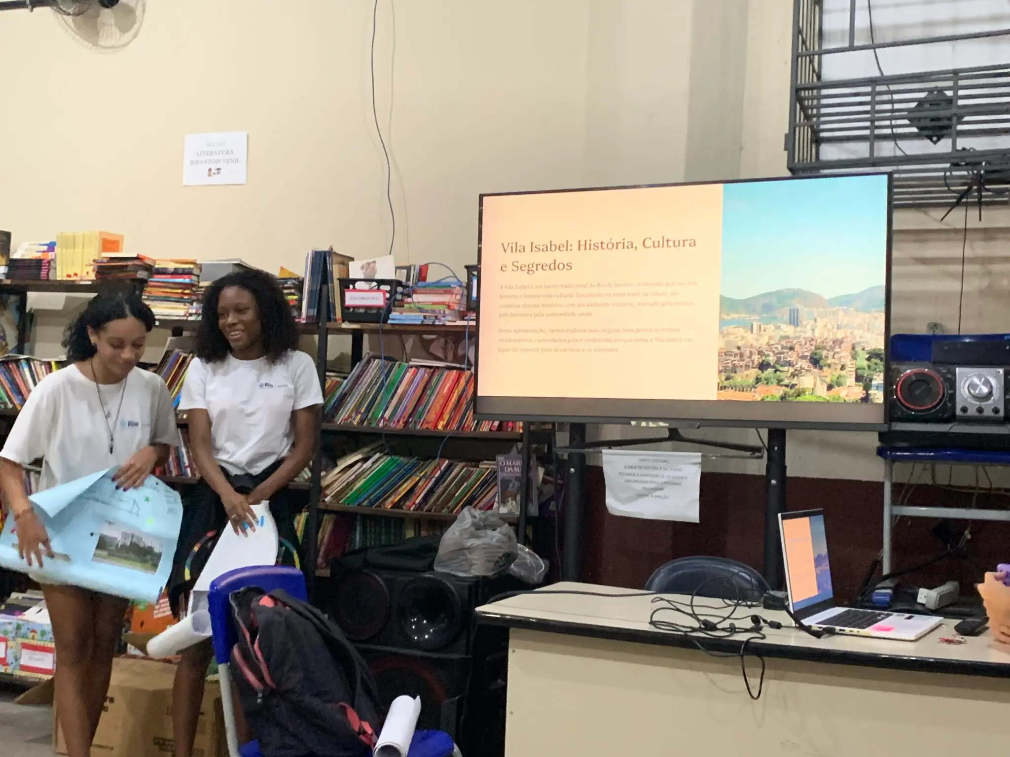 Duas mulheres em biblioteca com objetos e monitor exibindo "Vila Isabel: História, Cultura e Segredos".
