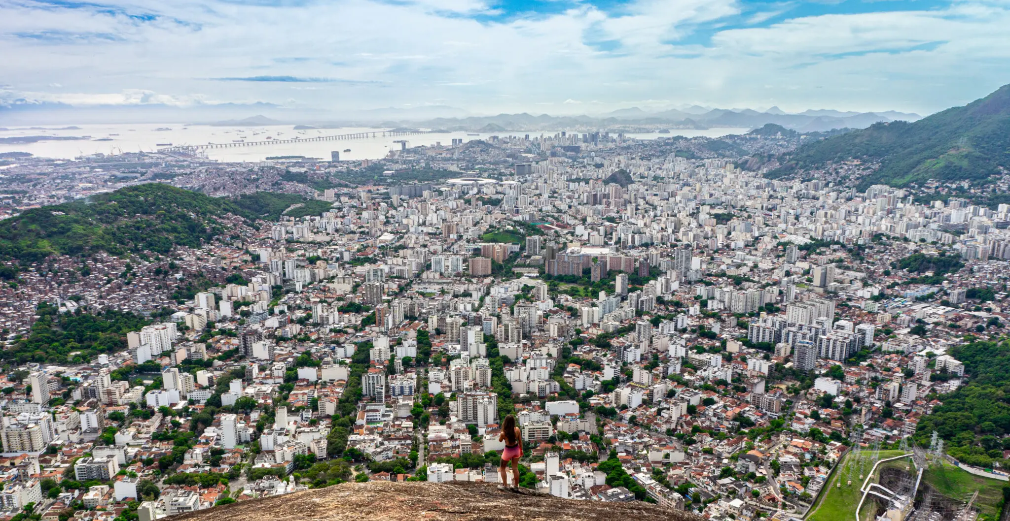 Cidade grande vista aérea com áreas verdes, edifícios, montanha, mar e pessoa em rocha.