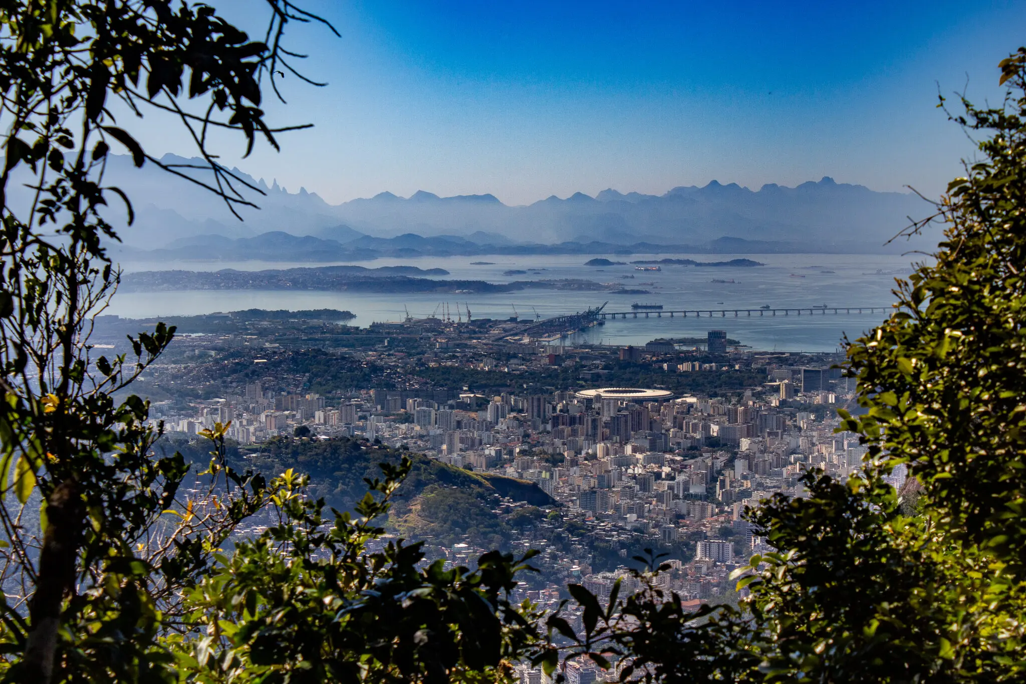 Cidade com vista panorâmica, vegetação, horizonte, edifícios, ponte, montanhas, céu azul, mar calmo.