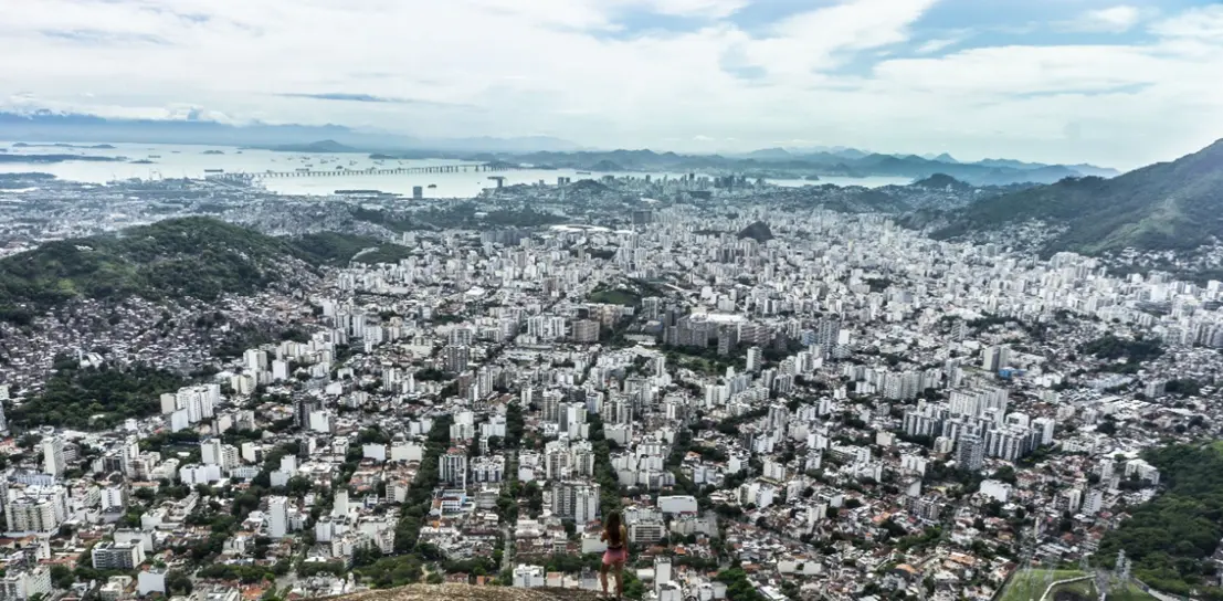 Cidade com vista panorâmica, áreas verdes, edifícios, montanhas, mar e céu nublado.