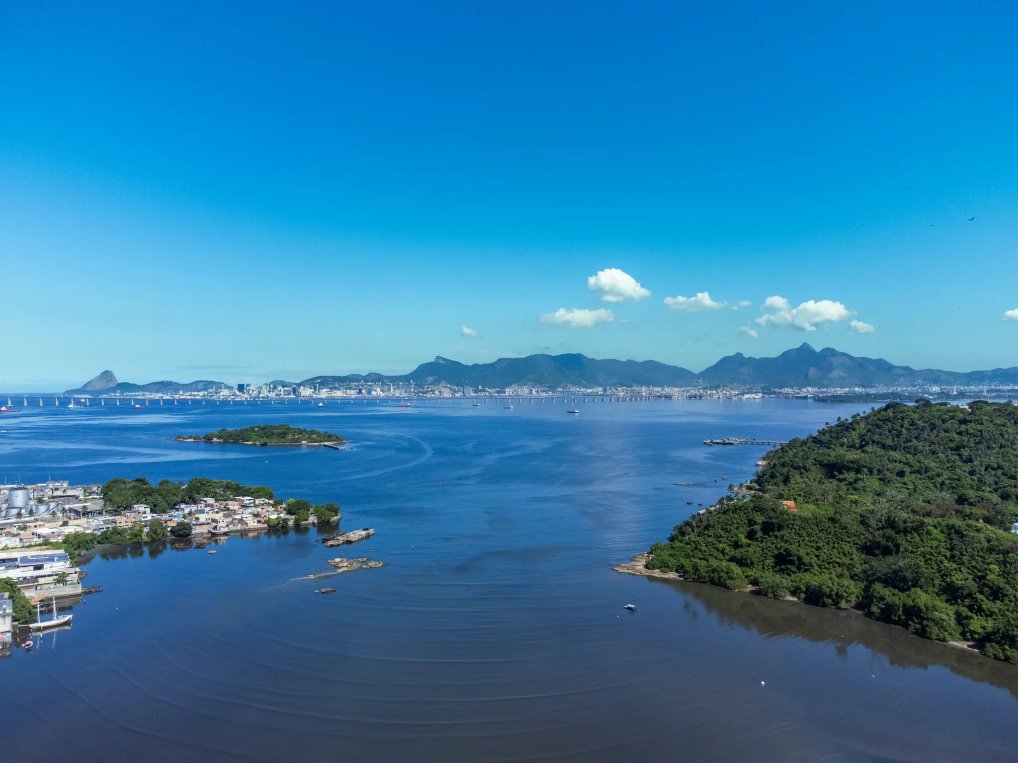 Paisagem costeira com céu azul, ilha verde e áreas urbanas e vegetadas.