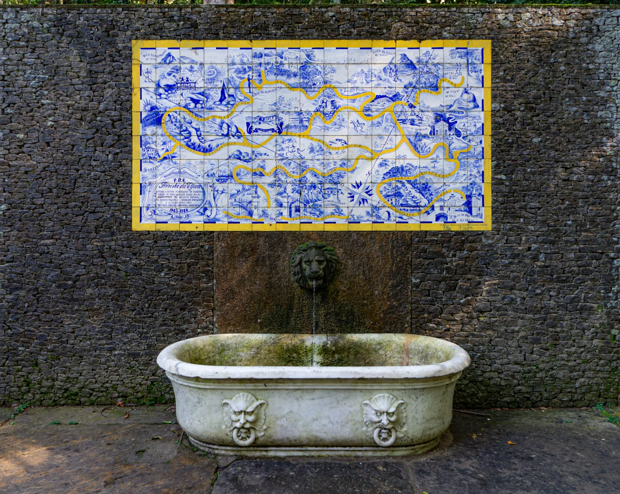Fonte com leão de pedra derramando água em tanque circular, parede de tijolos escuros e painel azul e branco.