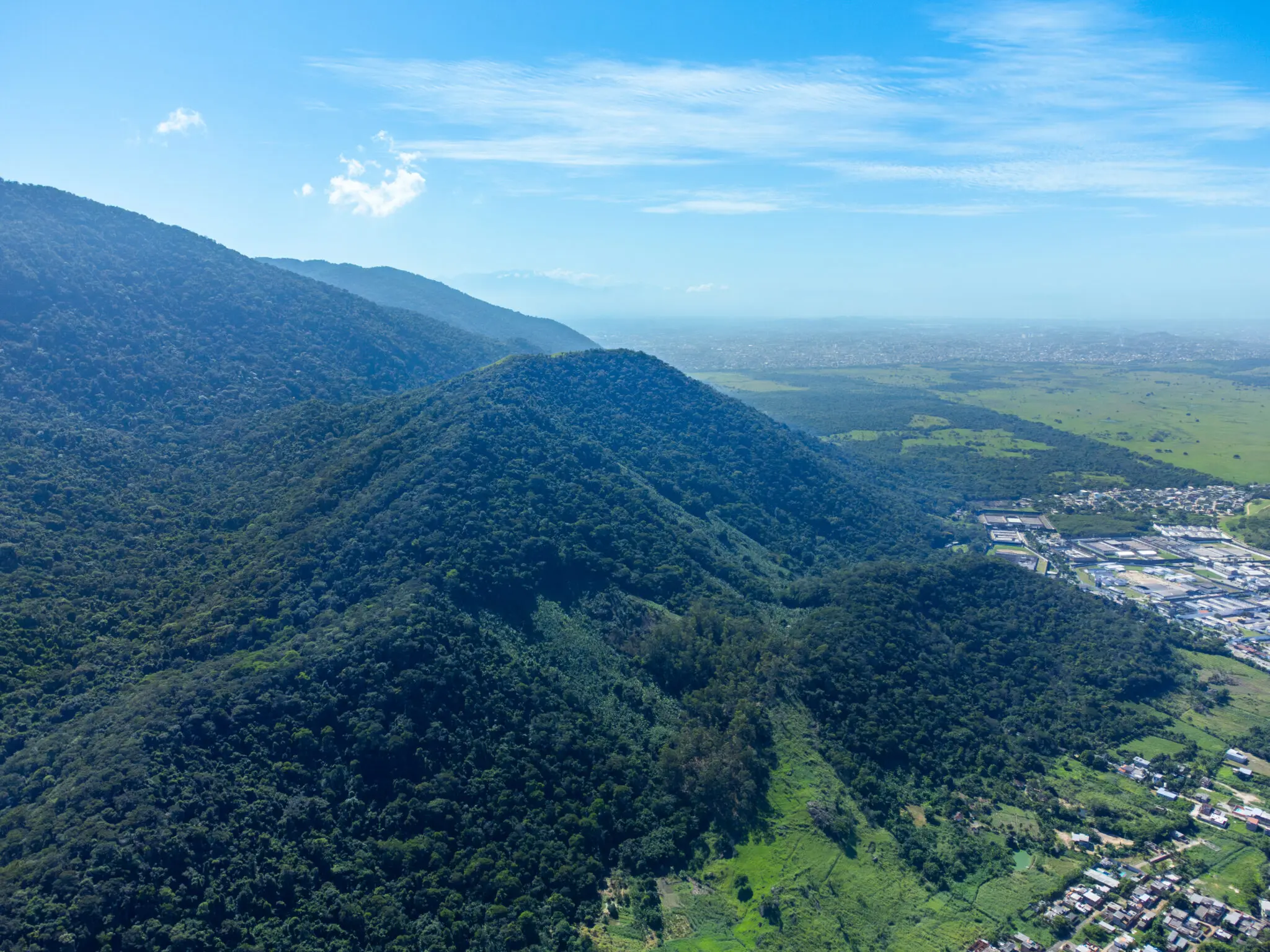 Maciço montanhoso com vegetação densa, colinas verdes e área plana com campos e construções.