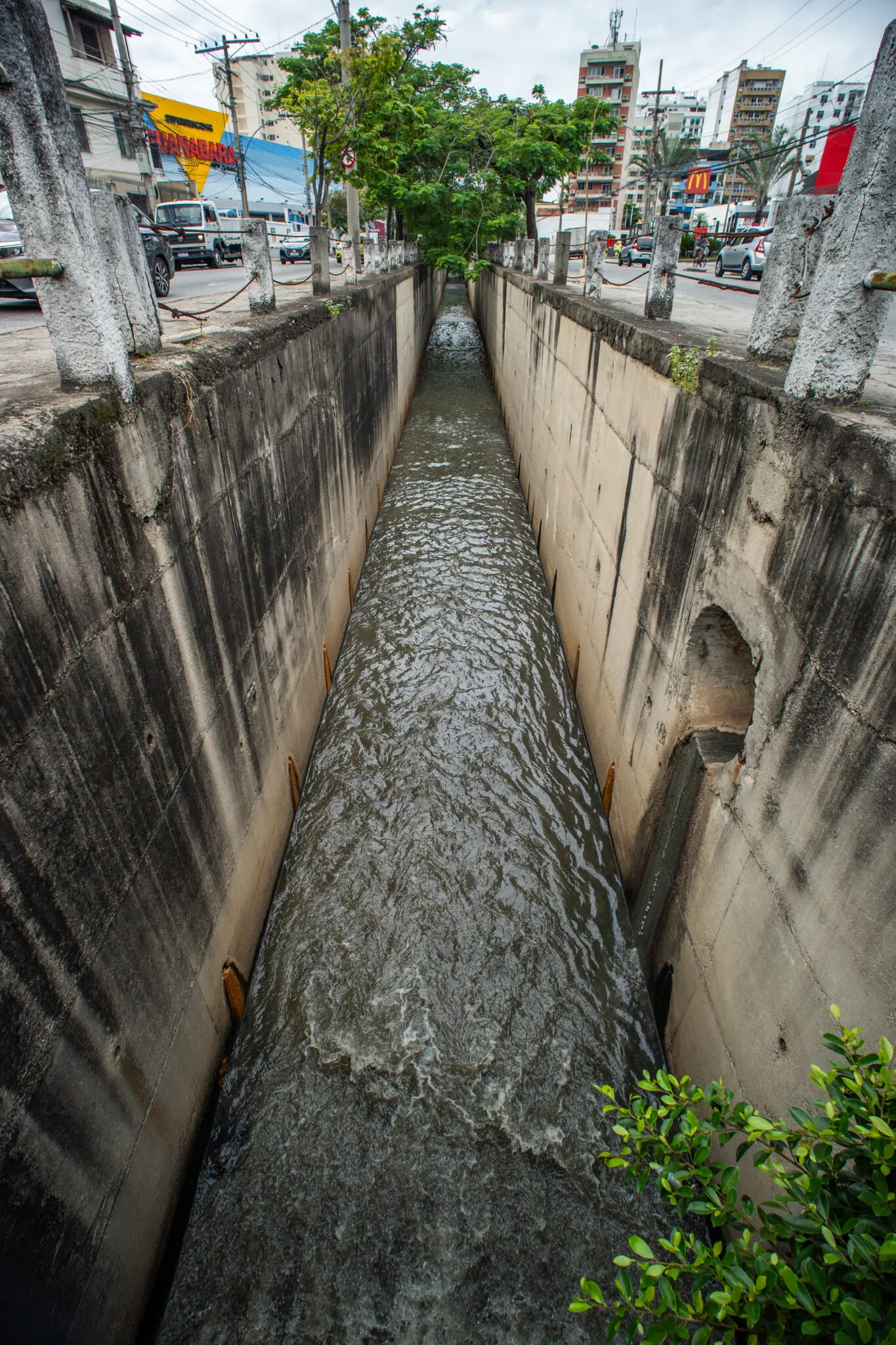 Canal de drenagem urbano com árvores e edifícios ao fundo.