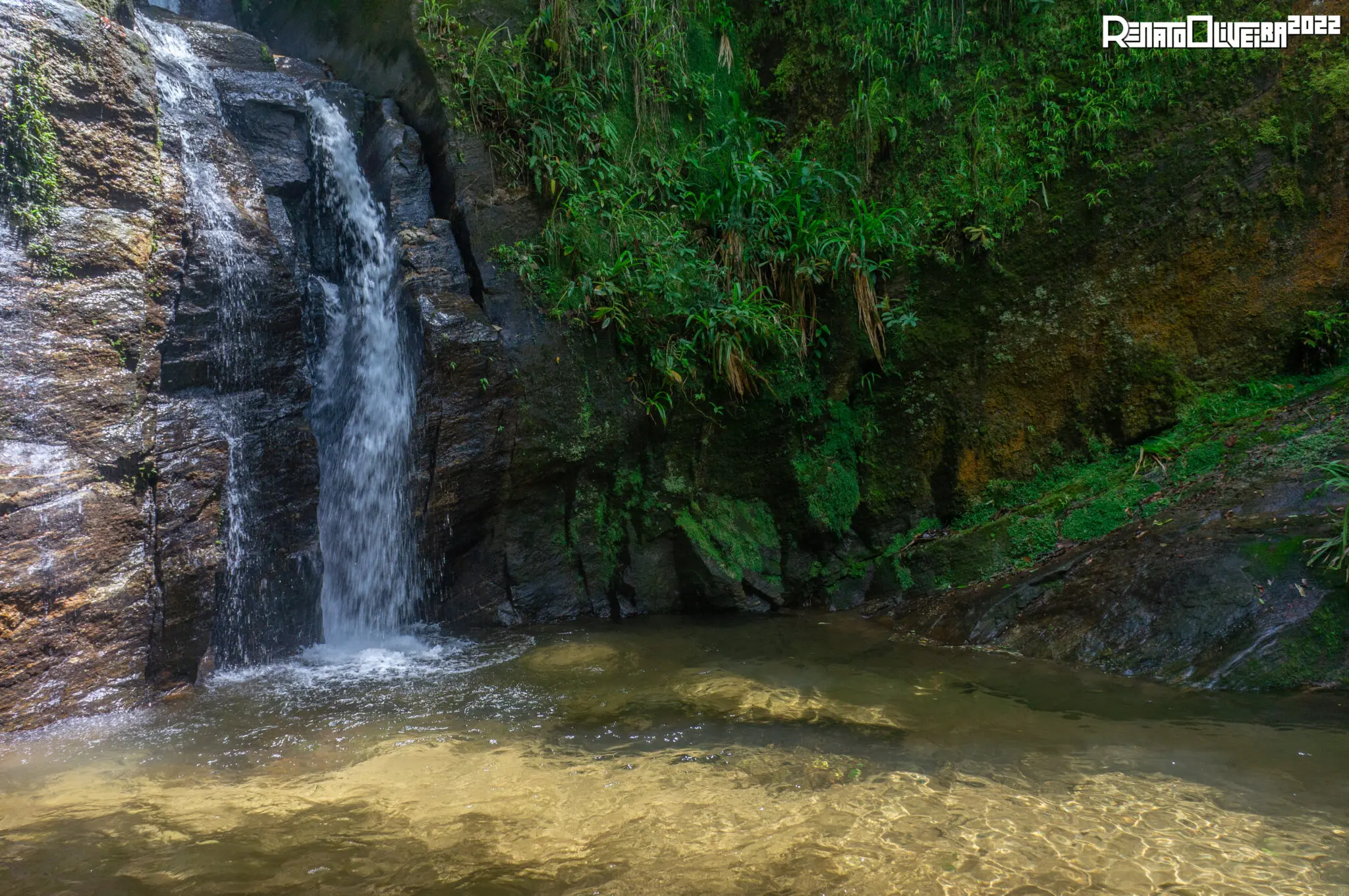 Cachoeira na Zona Sul, com vegetação verde e formação rochosa ao fundo.