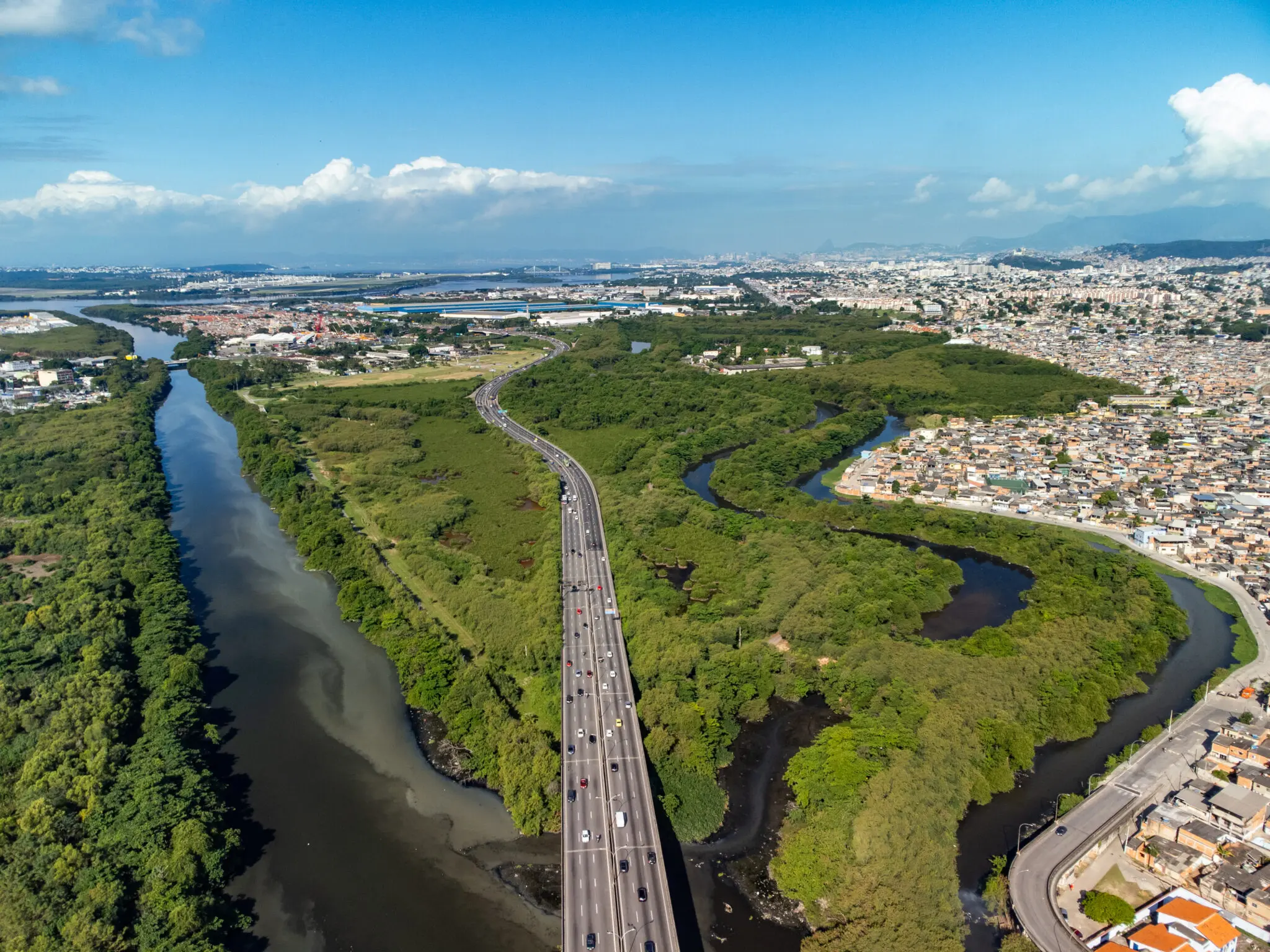 Área urbana e natural com rio, estrada, áreas verdes e cidade ao fundo.