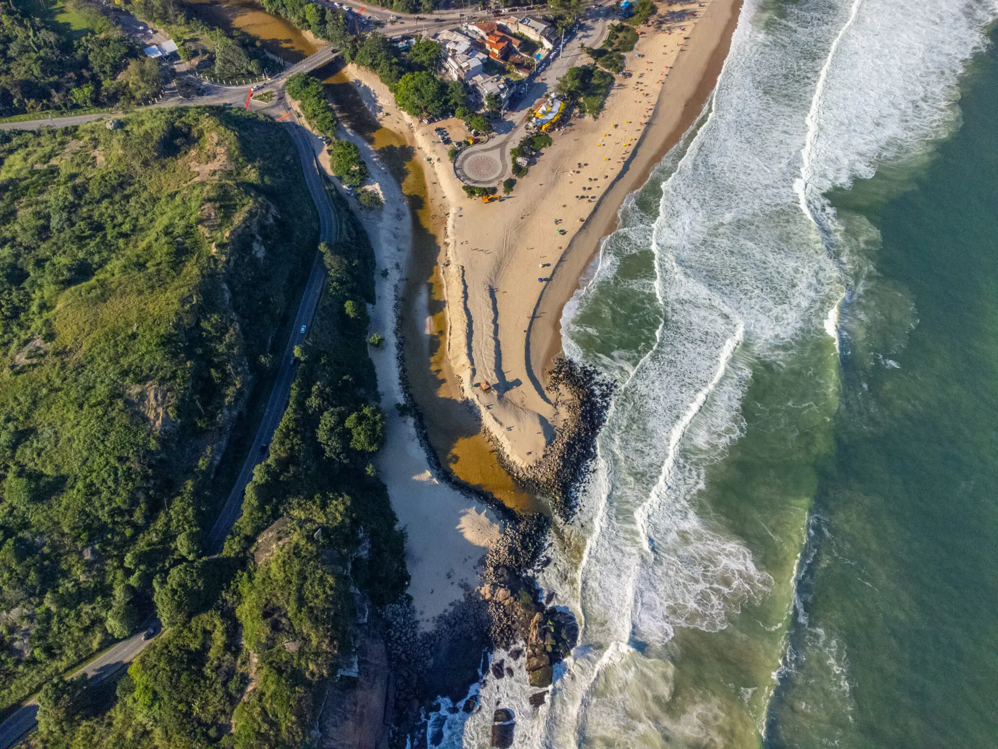 Praia costeira com estrada, vegetação verde, areia, construção e mar com ondas.