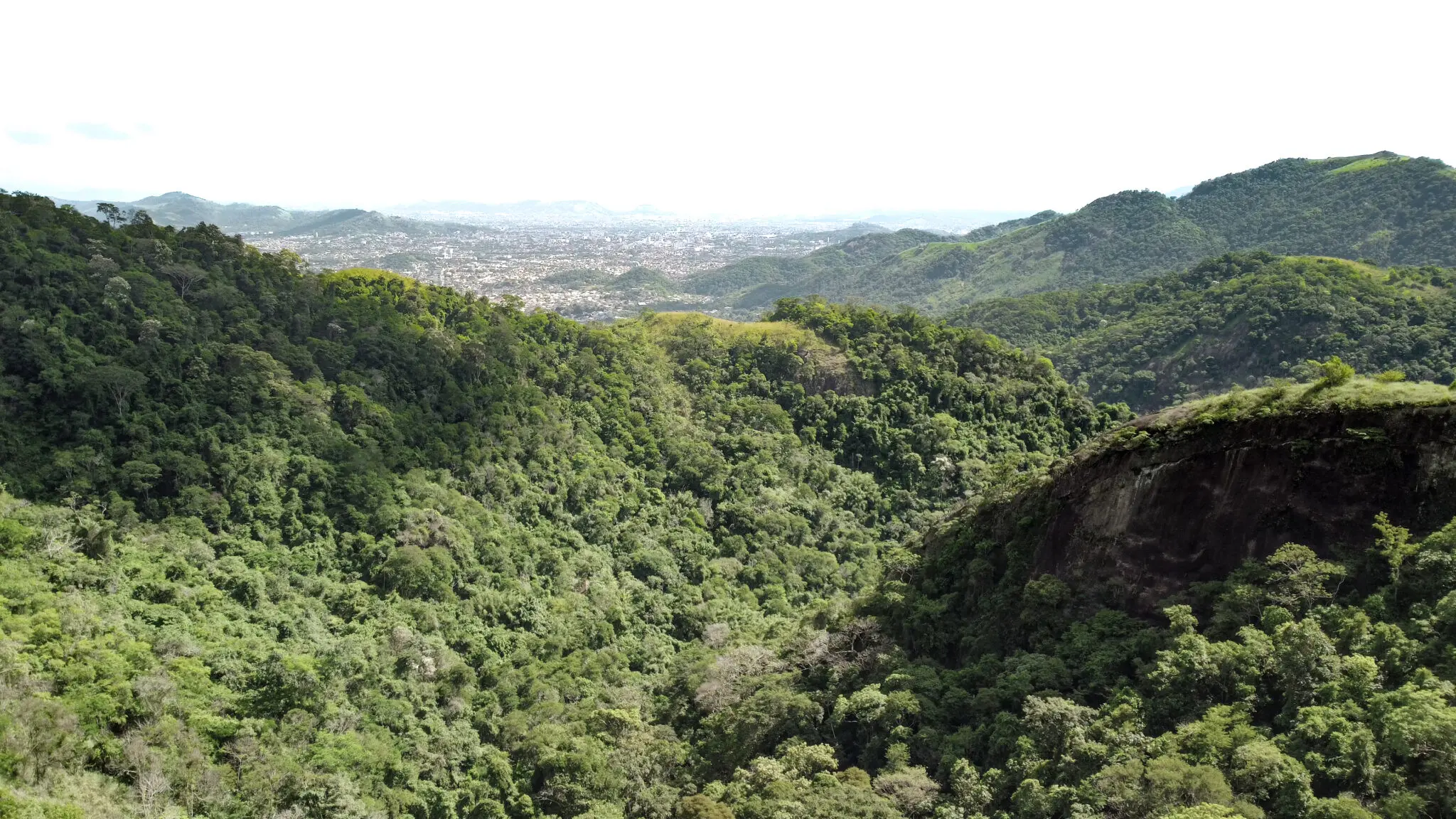 Paisagem montanhosa com colinas verdes, área urbana e céu azul.