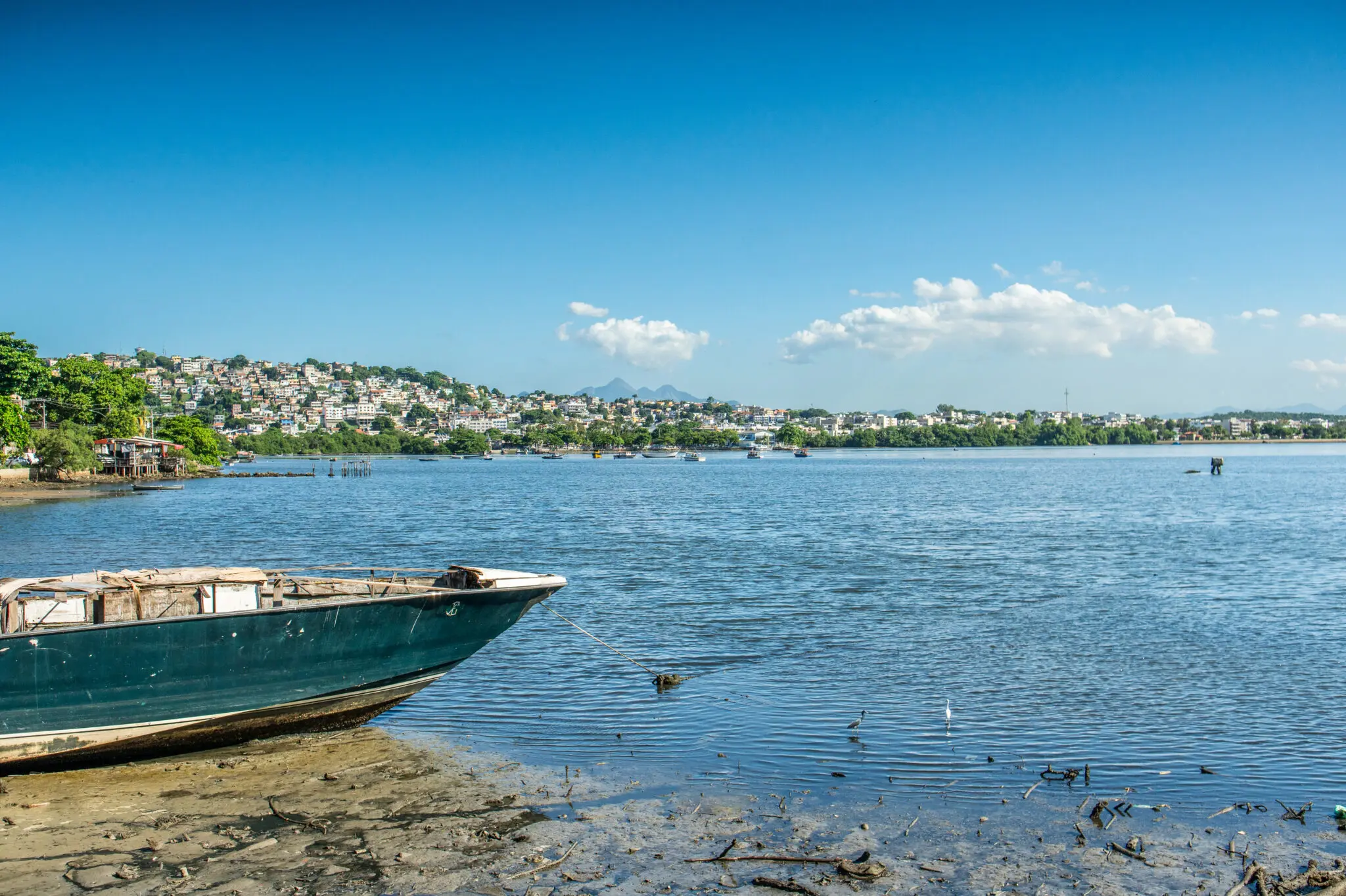 Praia com barco azul, cidade e montanhas ao fundo.