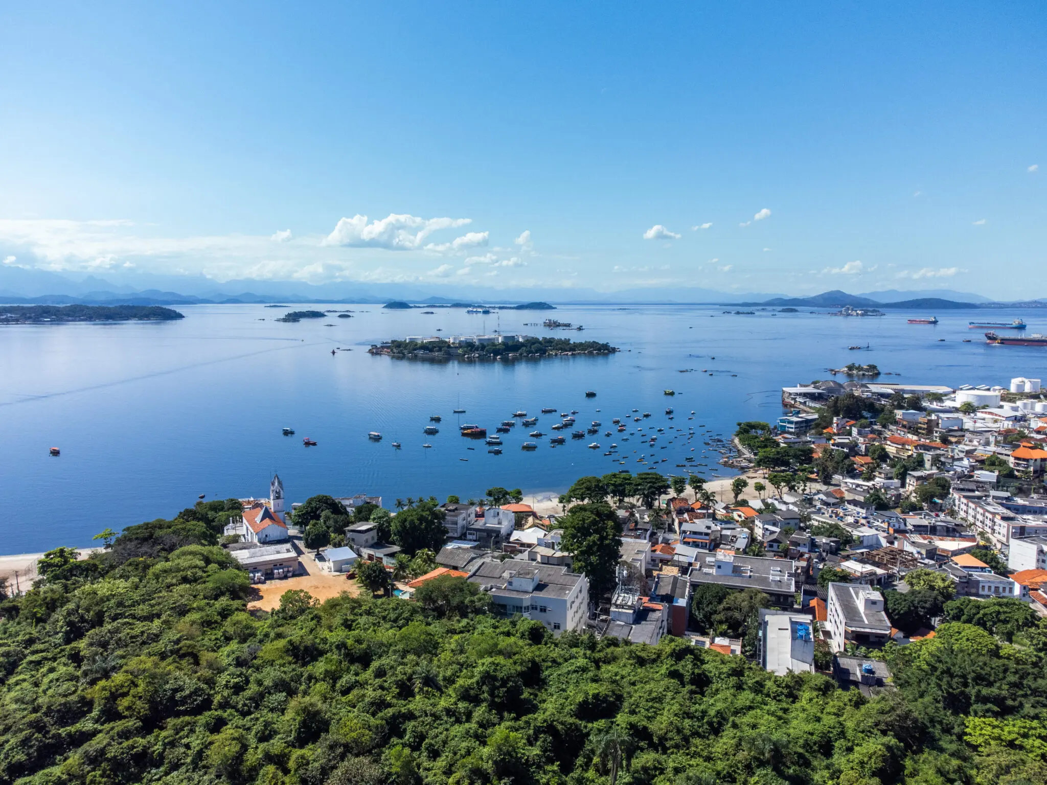 Praia da Baía de Guanabara com árvores, casas coloridas, água e montanhas.