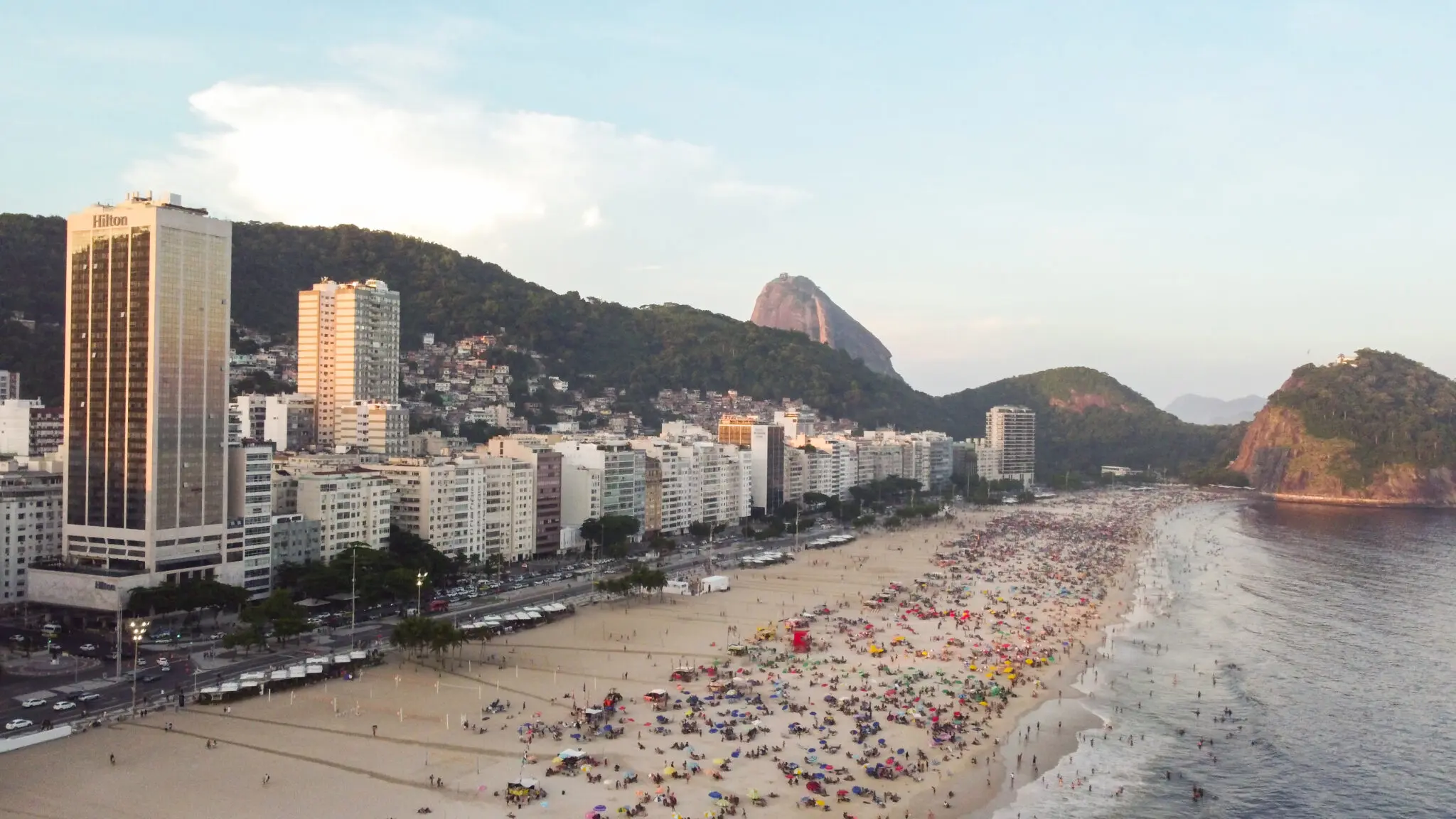 Praia lotada durante o dia com hotel Hilton e colina verde ao fundo.