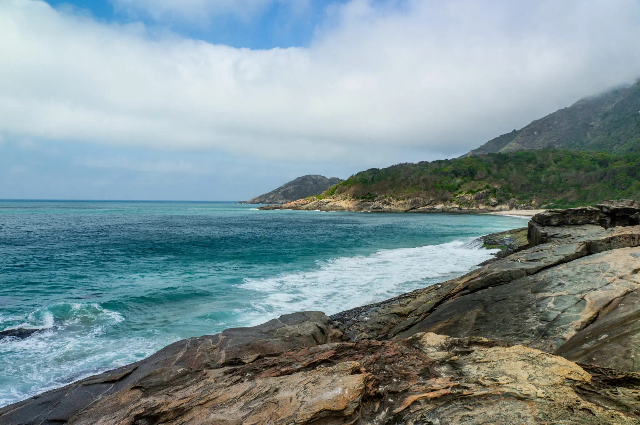 Praia costeira com rochas, vegetação verde e montanha ao fundo.