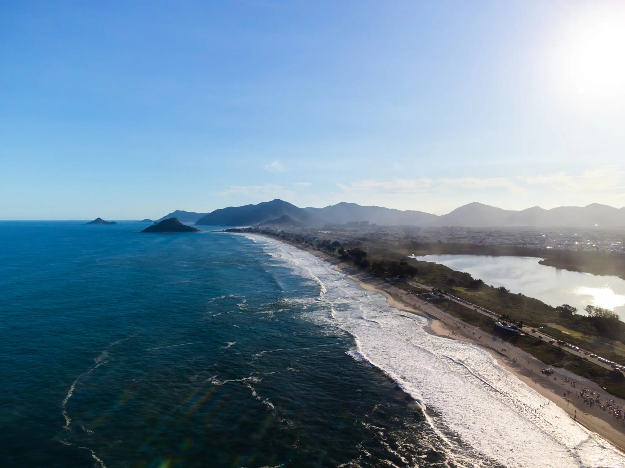 Praia costeira com oceano azul, areia bege, árvores verdes e cidade ao fundo.