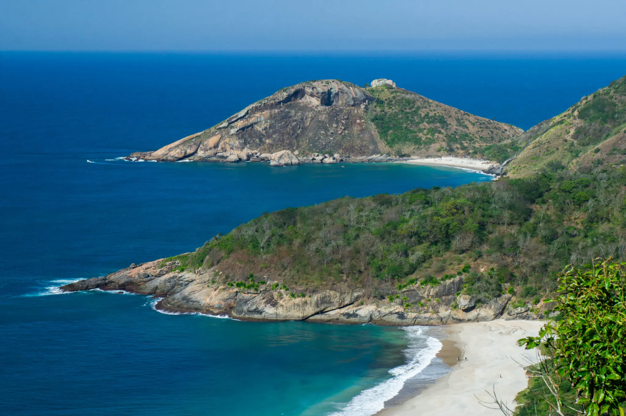Praia costeira com oceano azul, penhasco rochoso e vegetação densa.