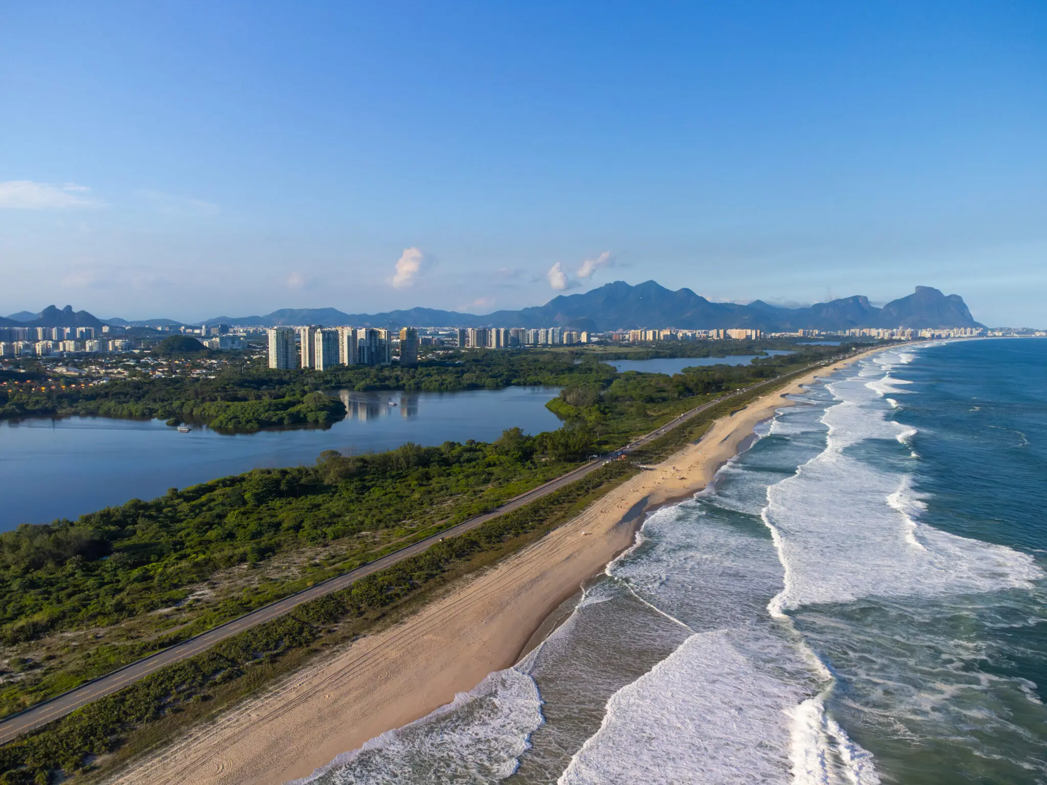 Praia costeira com vista panorâmica, mar em movimento, edifícios e montanhas ao fundo.