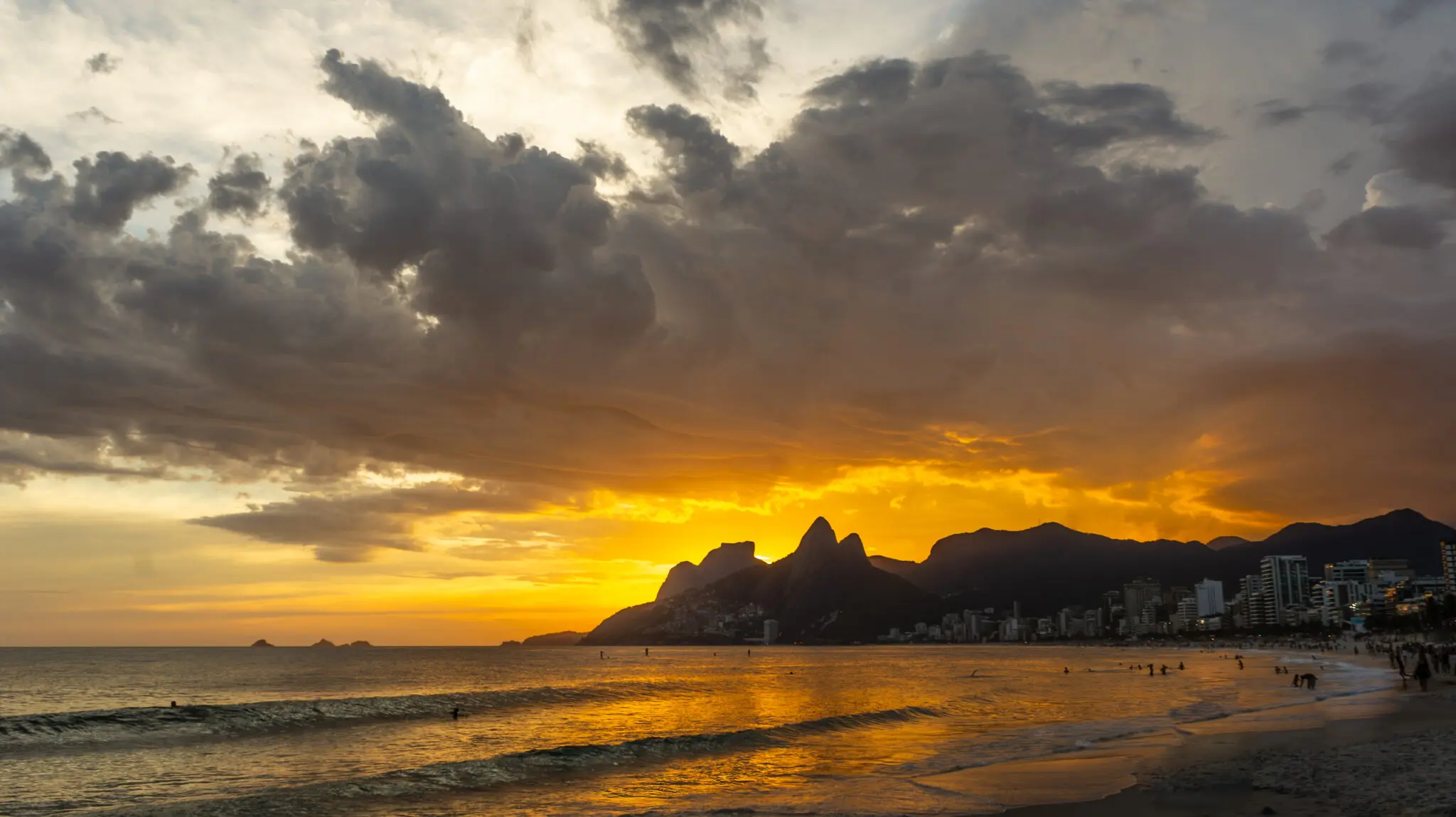 Praia durante pôr-do-sol com montanhas, prédios e pessoas desfrutando.