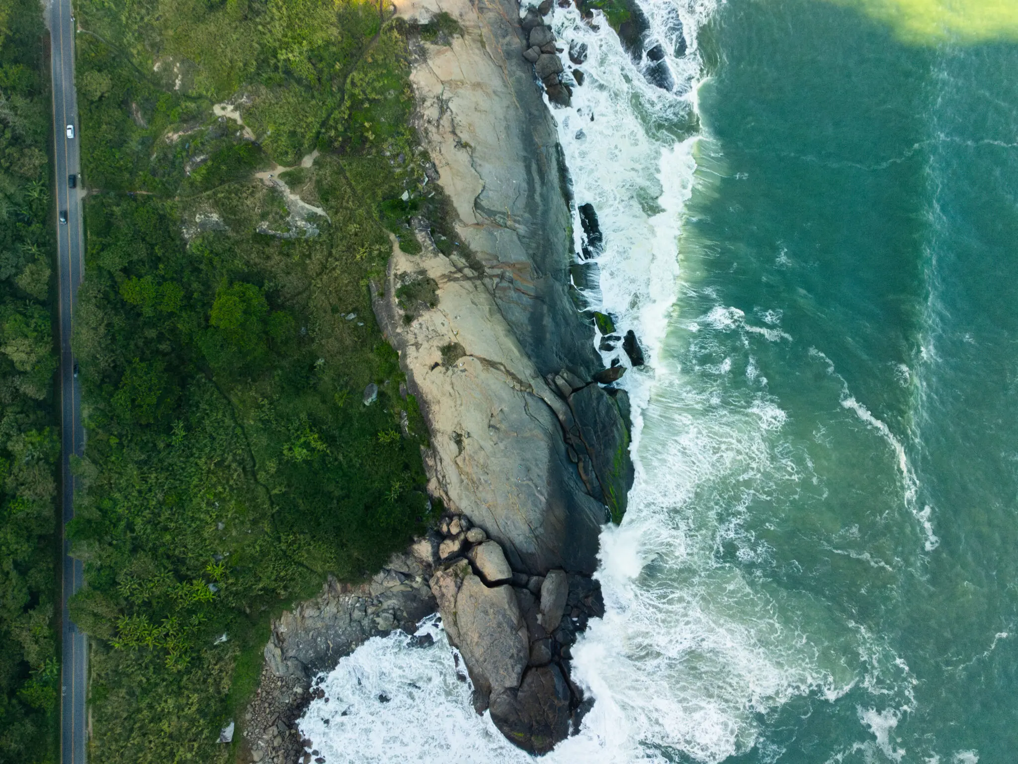 Praia costeira com estrada, vegetação, penhasco e mar em movimento.