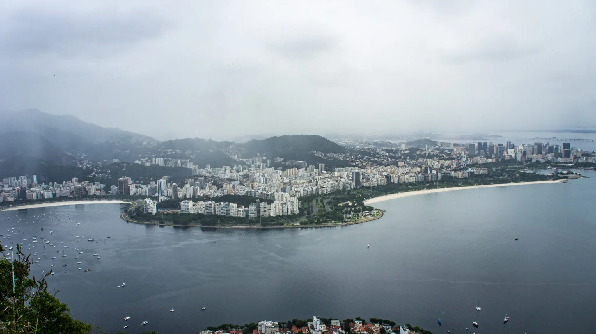 Cidade costeira sob céu nublado com praia, ilha moderna e montanhas verdes.