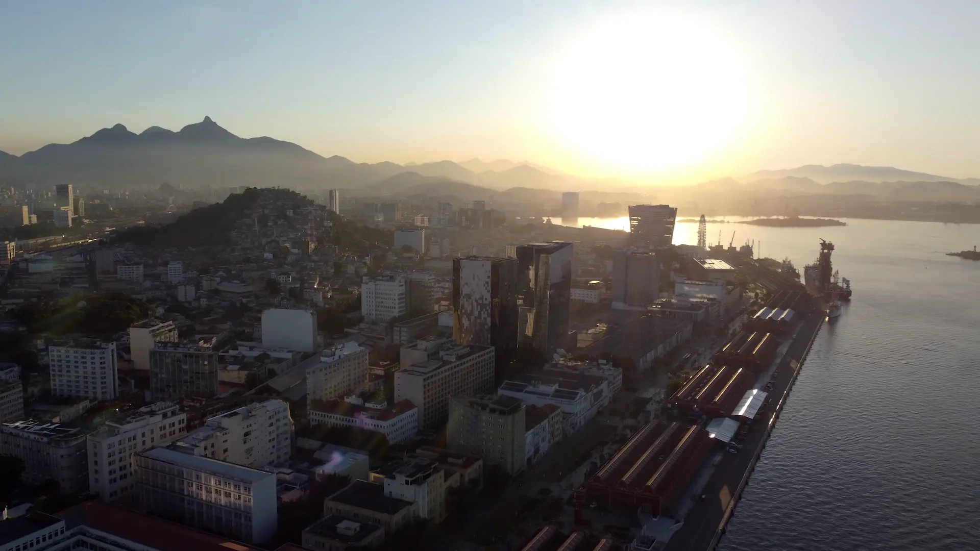 Cidade ao amanhecer com montanhas nebulosas e sol nascente.