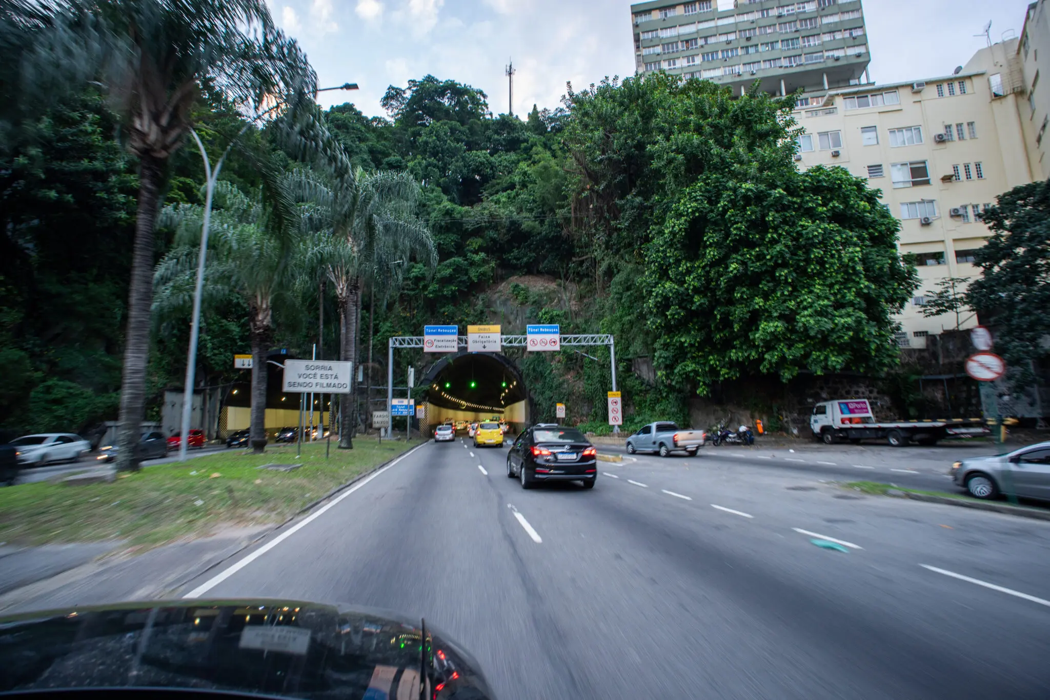 Estrada urbana com tráfego, árvores verdes, túnel com luzes verdes, edifícios residenciais altos, veículos em movimento e placa de sinalização de proibição de estacionamento.