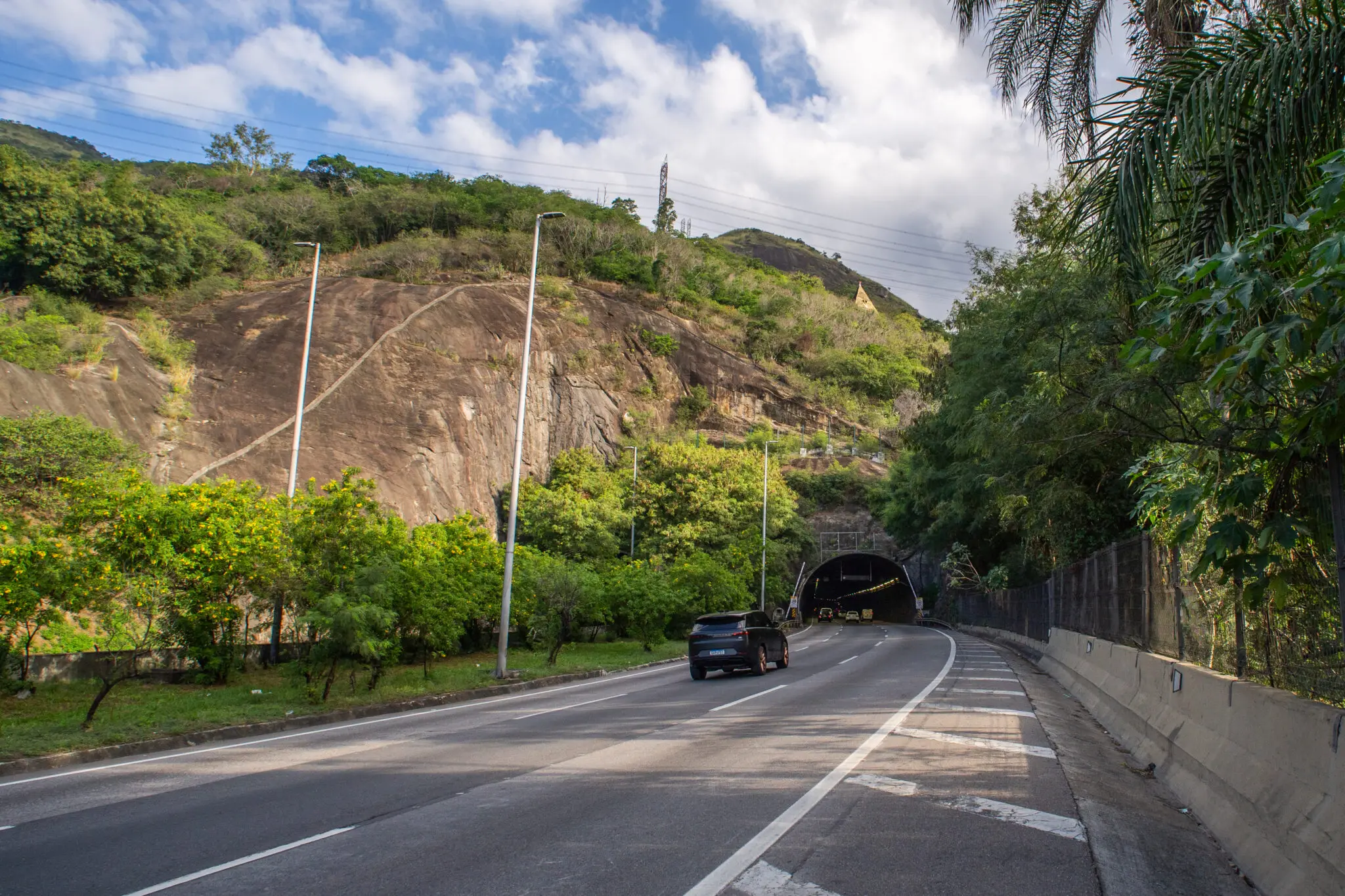 Estrada com túnel, colina verde, carro parado, barreira de concreto, céu azul com nuvens.