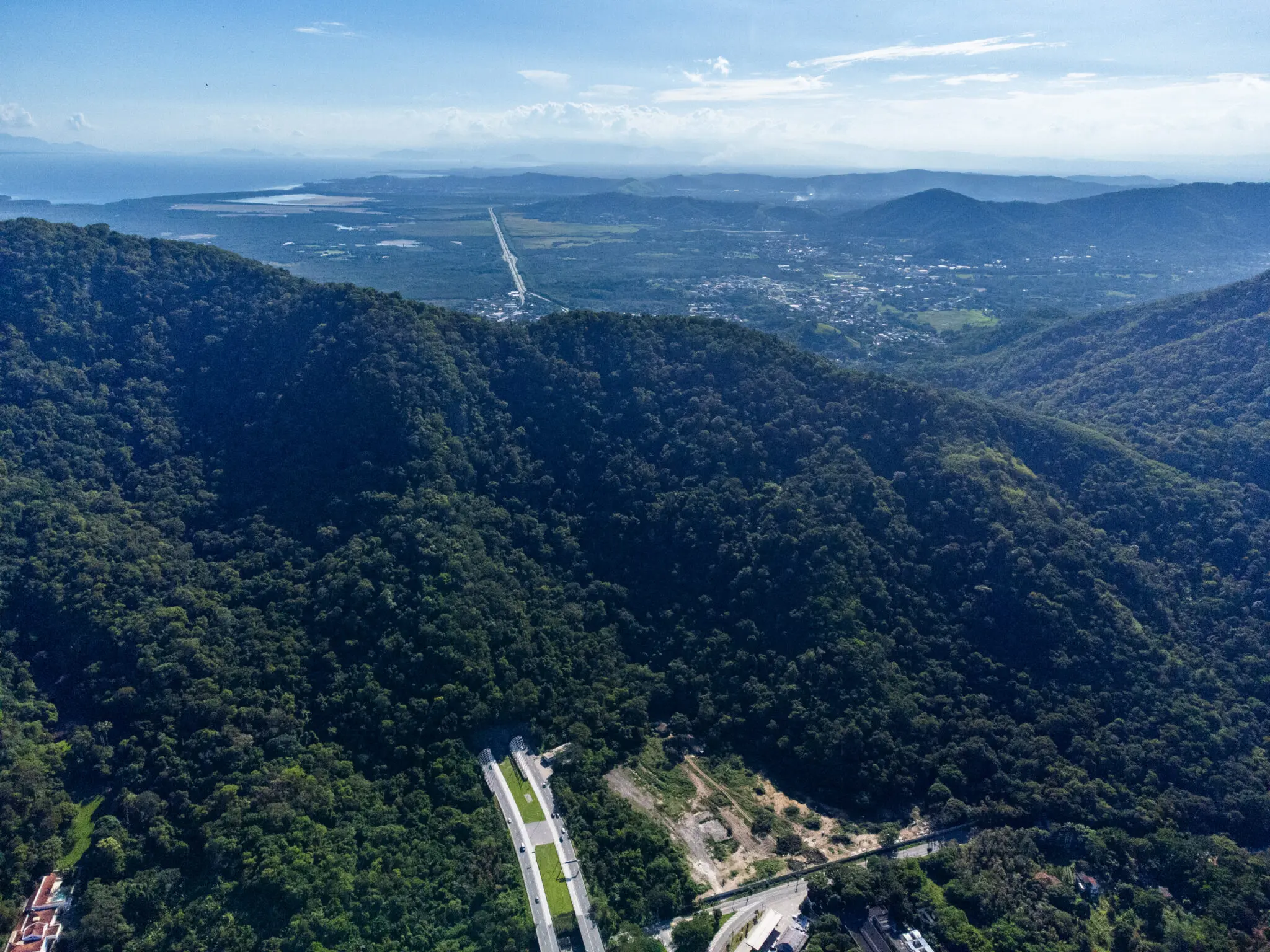 Área montanhosa com vegetação densa, estrada clara, planície aberta e água no horizonte.