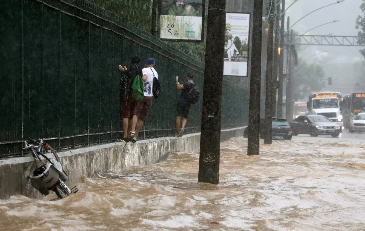 Uma rua inundada durante chuva forte, com pessoas observando e veículos parados.