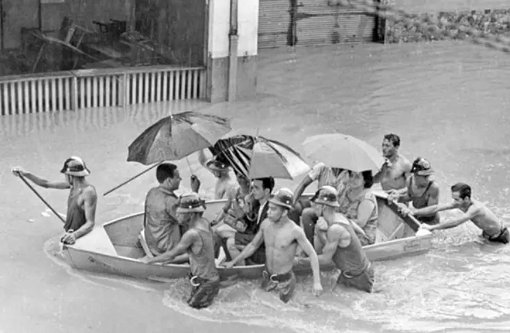 Inundações com pessoas em barca e prédio submerso.