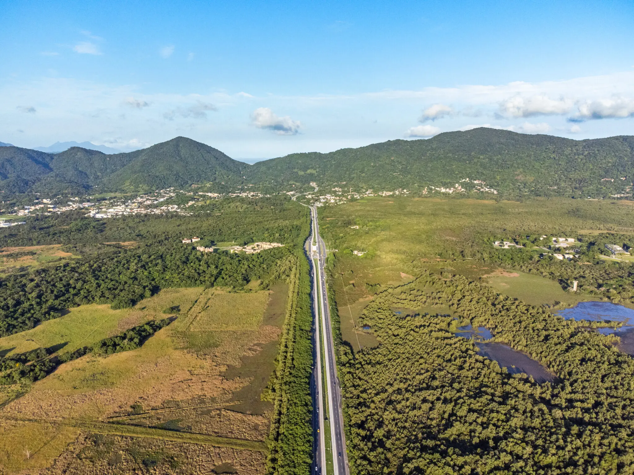 Área rural com estrada centralizada, campos agrícolas, florestas, áreas verdes, lago e montanhas cobertas de vegetação.