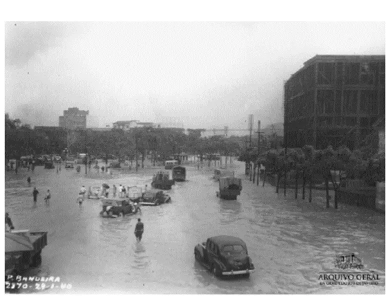 Rua inundada com pessoas caminhando e veículos tentando atravessar água.