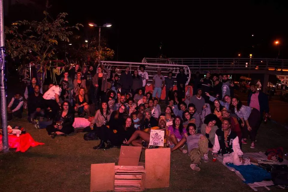 Grupo de pessoas reunidos em local noturno, sentados no chão e em caixas de madeira.