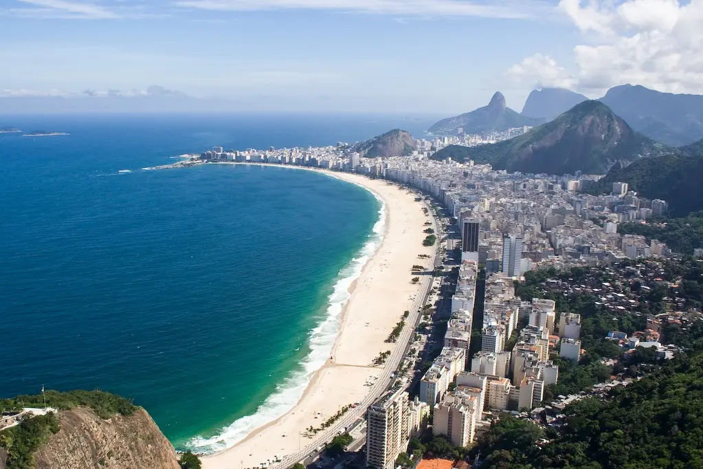 Praia costeira com cidade ao fundo, oceano azul e montanhas verdes.