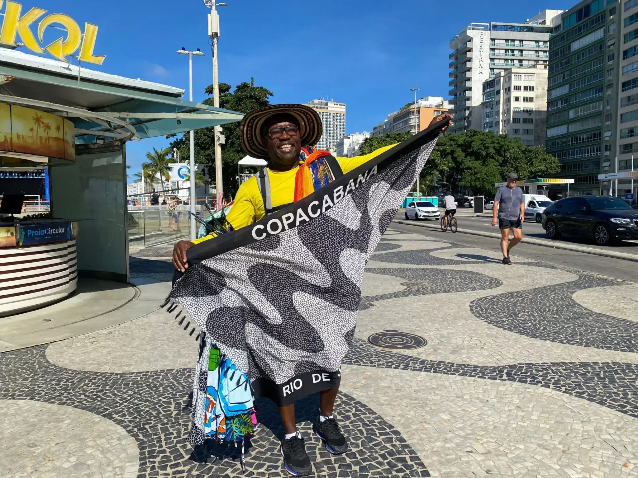 Homem sorridente com chapéu de palha e camisa amarela segurando lenço "COPACABANA" e "RIO DE", em local com pavimentação de pedras.