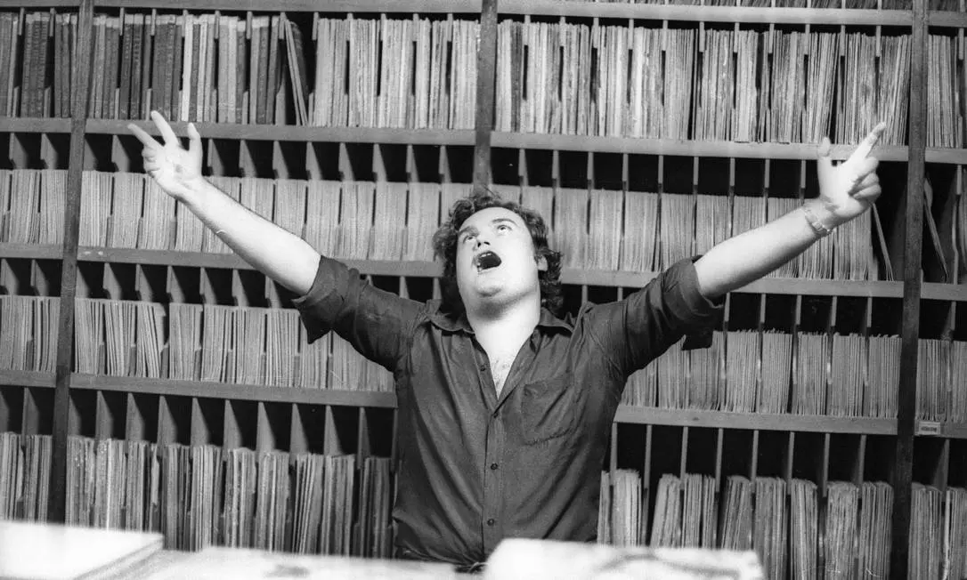 Homem em biblioteca, braços estendidos, olhando para cima.