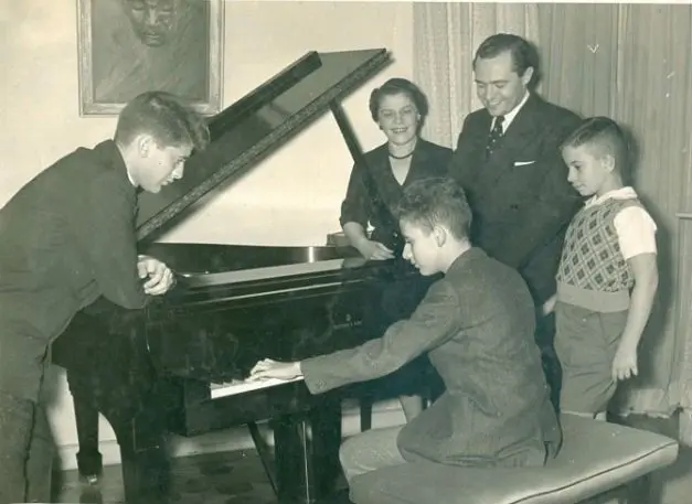 Família reunida ao redor de um piano, homem tocando, dois meninos assistindo, adultos sorrindo.
