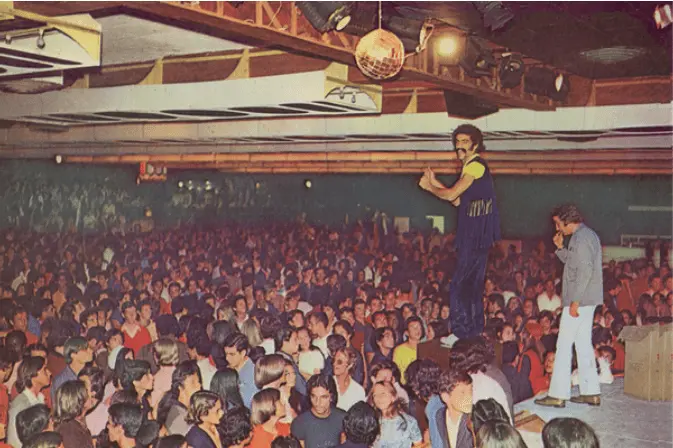 Baile de dança dos anos 1970, multidão dançando em salão amplo.