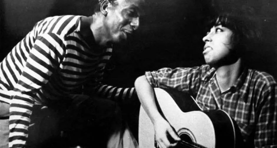 Dois amigos em cena íntima, trocando olhares com violão.