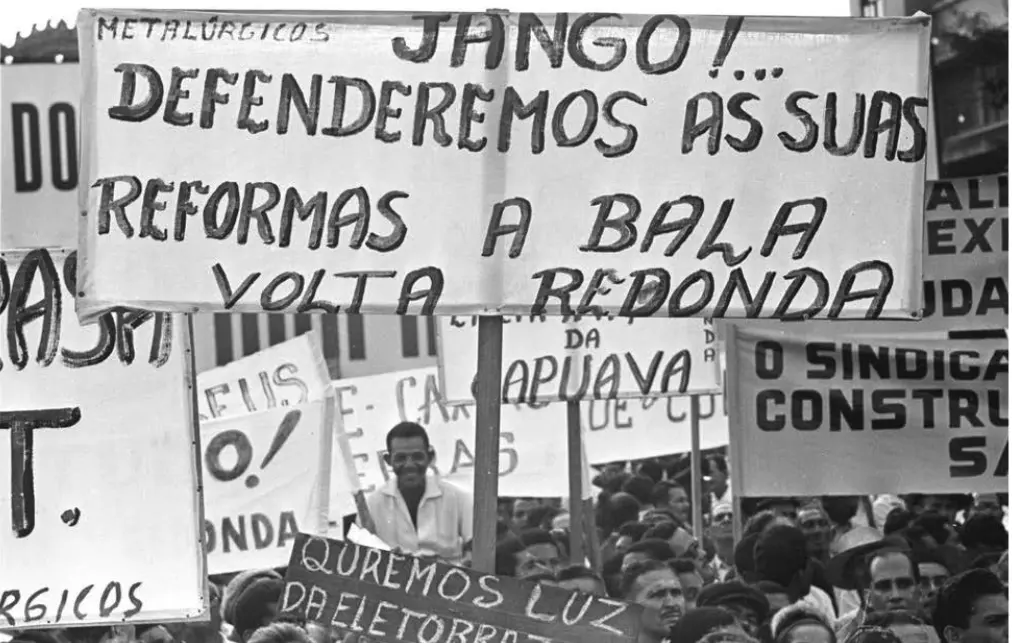 Multidão em comício com cartazes, homem sorridente no centro.
