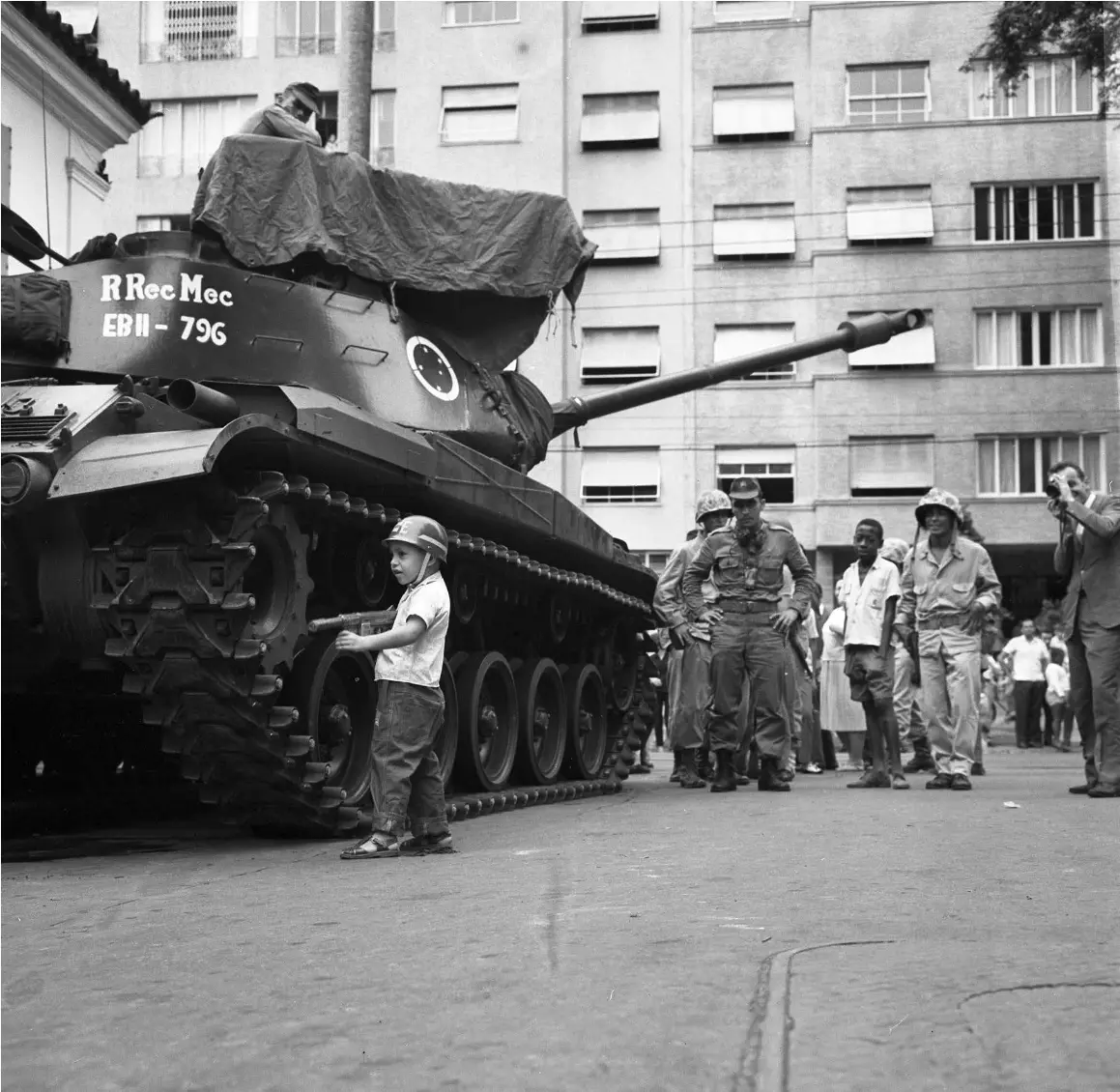 Tanque militar em cena urbana, menino com capacete, soldados e edifício ao fundo.