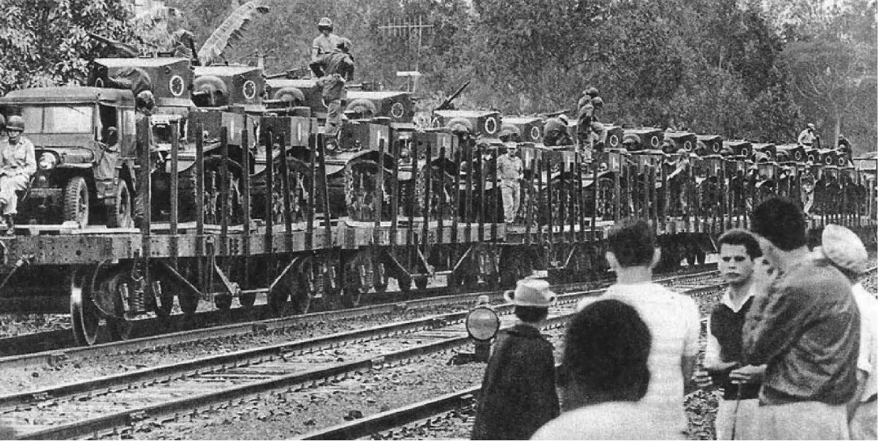 Formação militar com veículos e observadores civis em trilhas ferroviárias.
