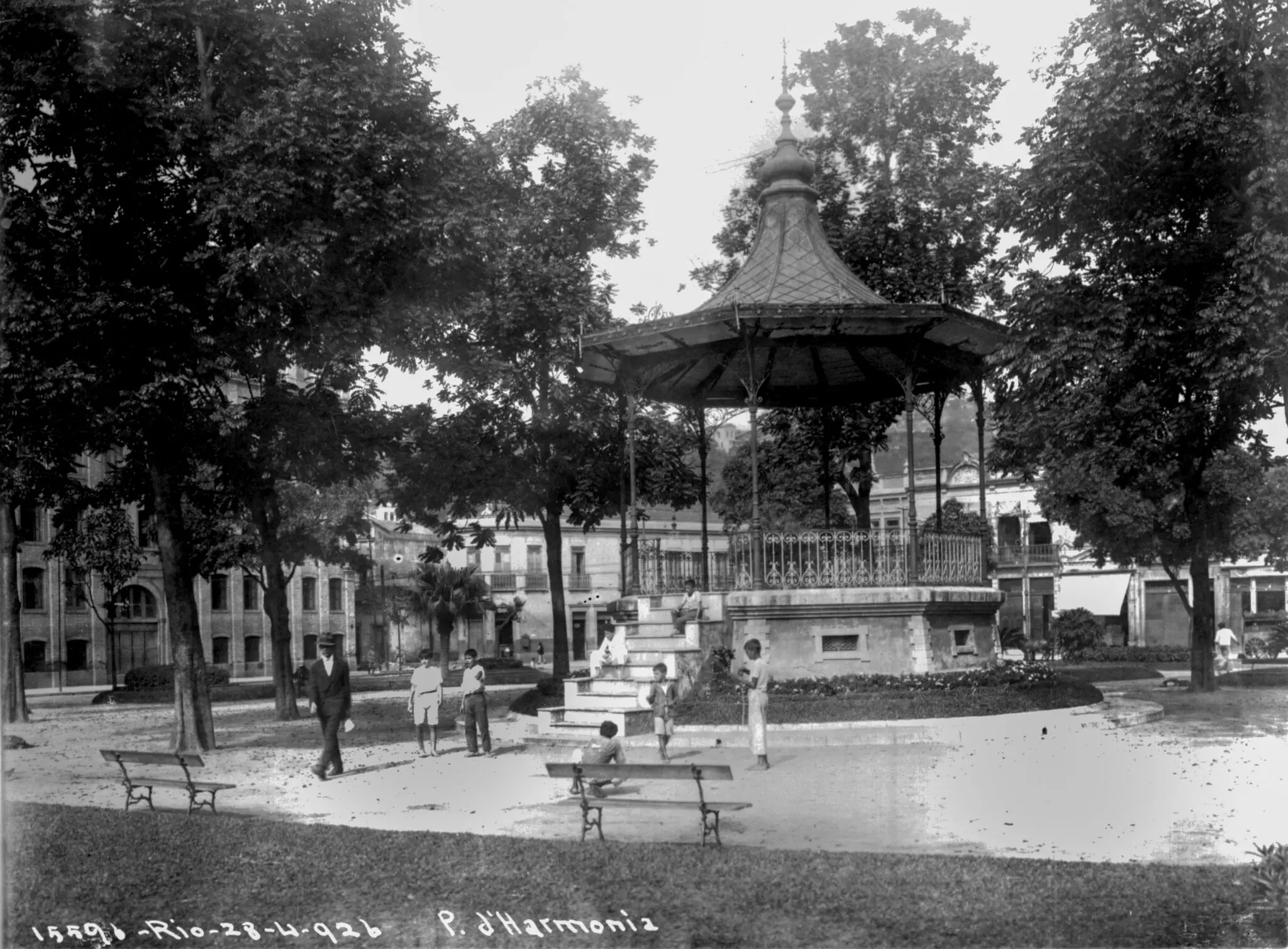 Praça antiga com árvores, kiosque metálico, pessoas caminhando e edifícios históricos.