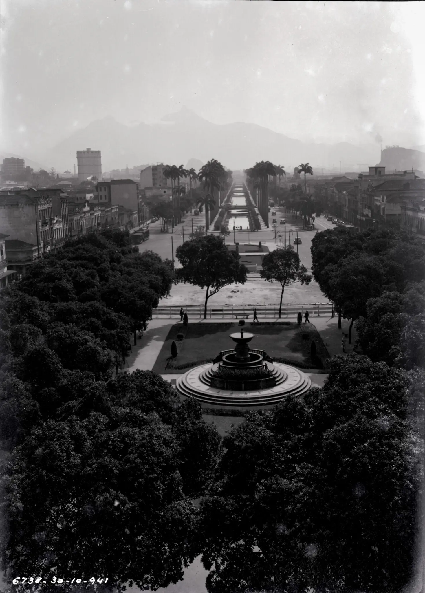 Praça com fonte, árvores, edifícios e alameda de palmeiras.