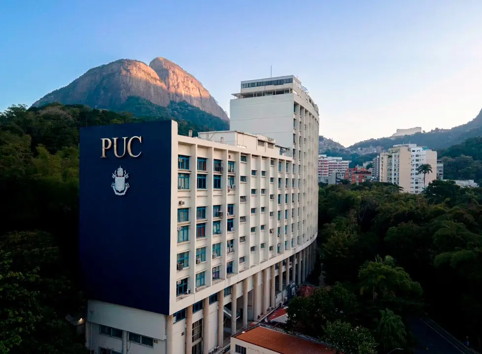 Prédio universitário PUC com estrutura curva e janelas, montanha ao fundo, céu azul claro.
